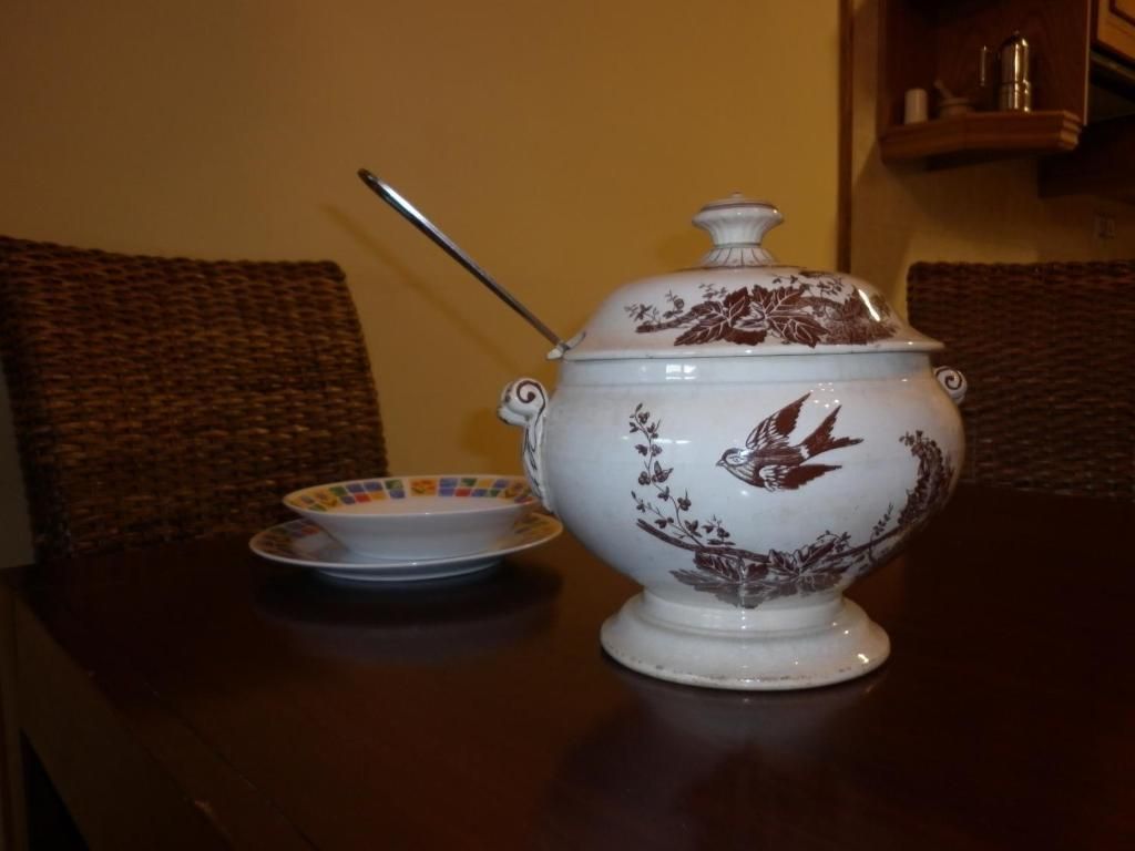 Ternera blanca amb disseny de paisatge i ocell marró, cullerot, plats i cadires de vímet sobre una taula fosca.