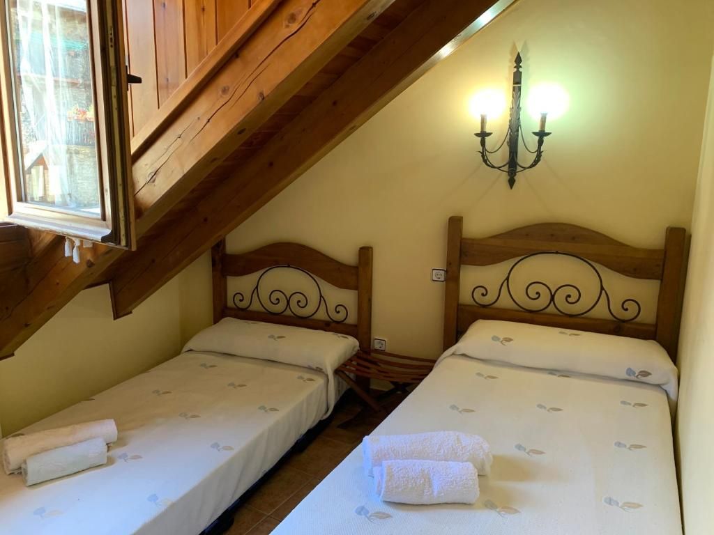 Dos llits individuals en una habitació amb sostre inclinat i finestra, petit aplic decoratiu a la paret.