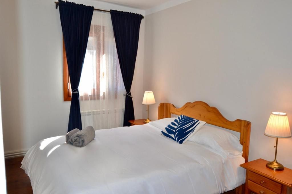 Dormitori amb un llit doble, roba de llit blanca, tauletes de nit, làmpades, finestra amb cortines blaves.