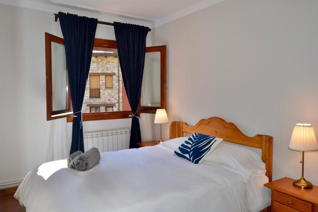 Dormitori amb un llit fet, tauletes de nit, una finestra amb cortines i una tovallola.