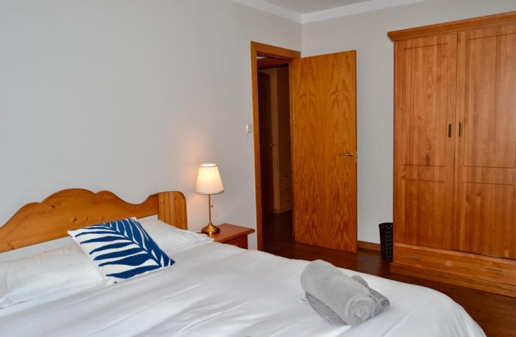 Dormitori amb llit, làmpada, armari, porta oberta al passadís. Paret grisa, mobles de fusta, roba de llit blanca.