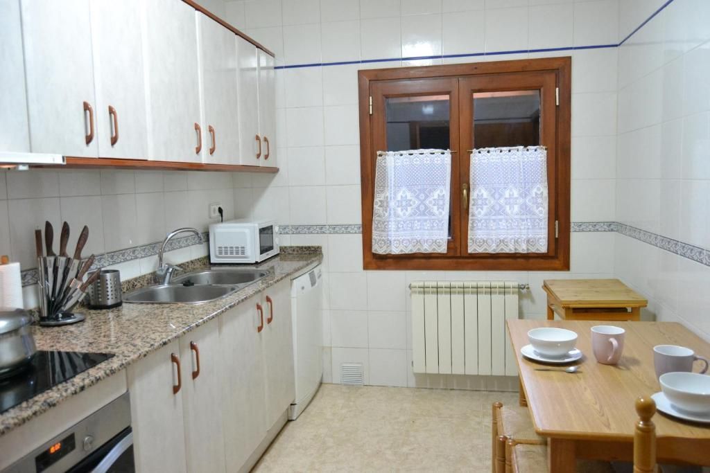 Cuina amb armaris blancs, taulell de granit, finestra amb cortines i una petita taula de menjador parada per a un àpat.