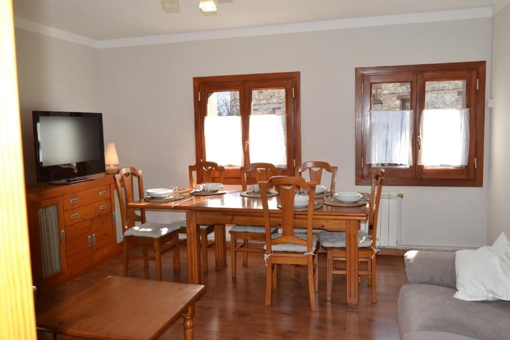 Menjador amb taula i cadires de fusta, finestres amb cortines, televisor i armaris marrons.