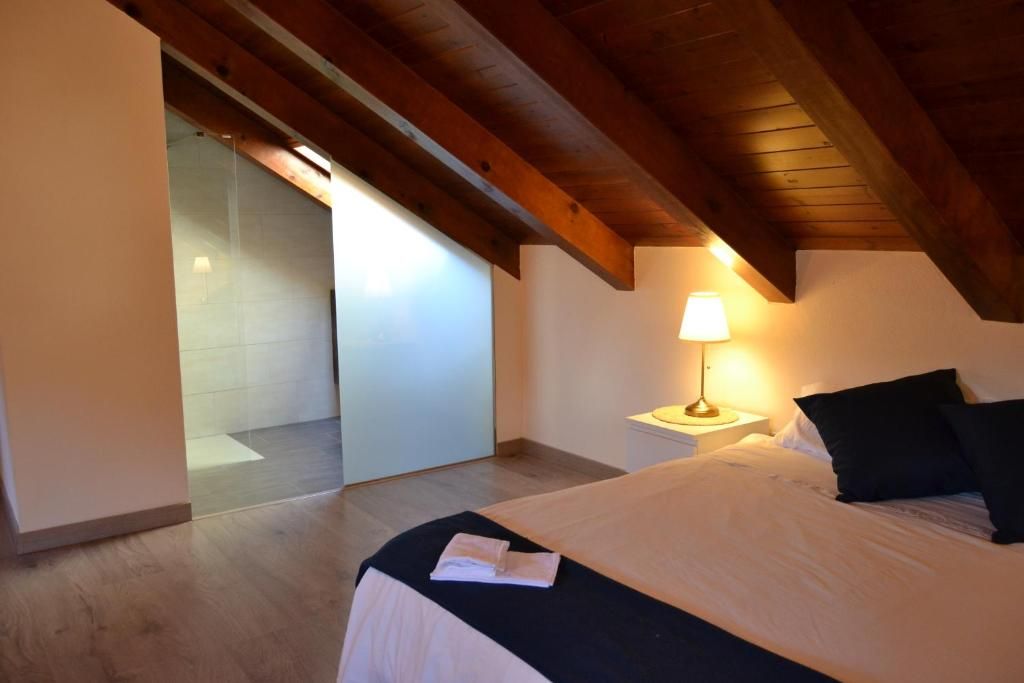 Dormitori amb bigues de fusta, llit, làmpada i porta de vidre esmerilat a una zona de dutxa.