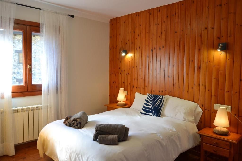 Dormitori amb paret amb panells de fusta, llit doble amb roba de llit blanca, tauletes de nit petites i finestra amb cortines transparents.