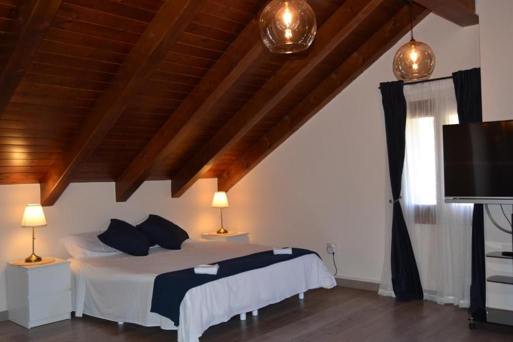Dormitori amb llit blanc, detalls blau fosc, sostre de fusta i dos llums penjants.