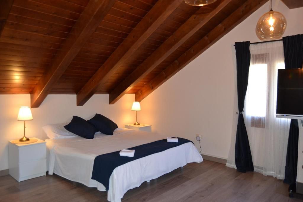 Dormitori amb sostre de bigues de fusta, llit king size, tauletes de nit, làmpades i una finestra amb cortines fosques.