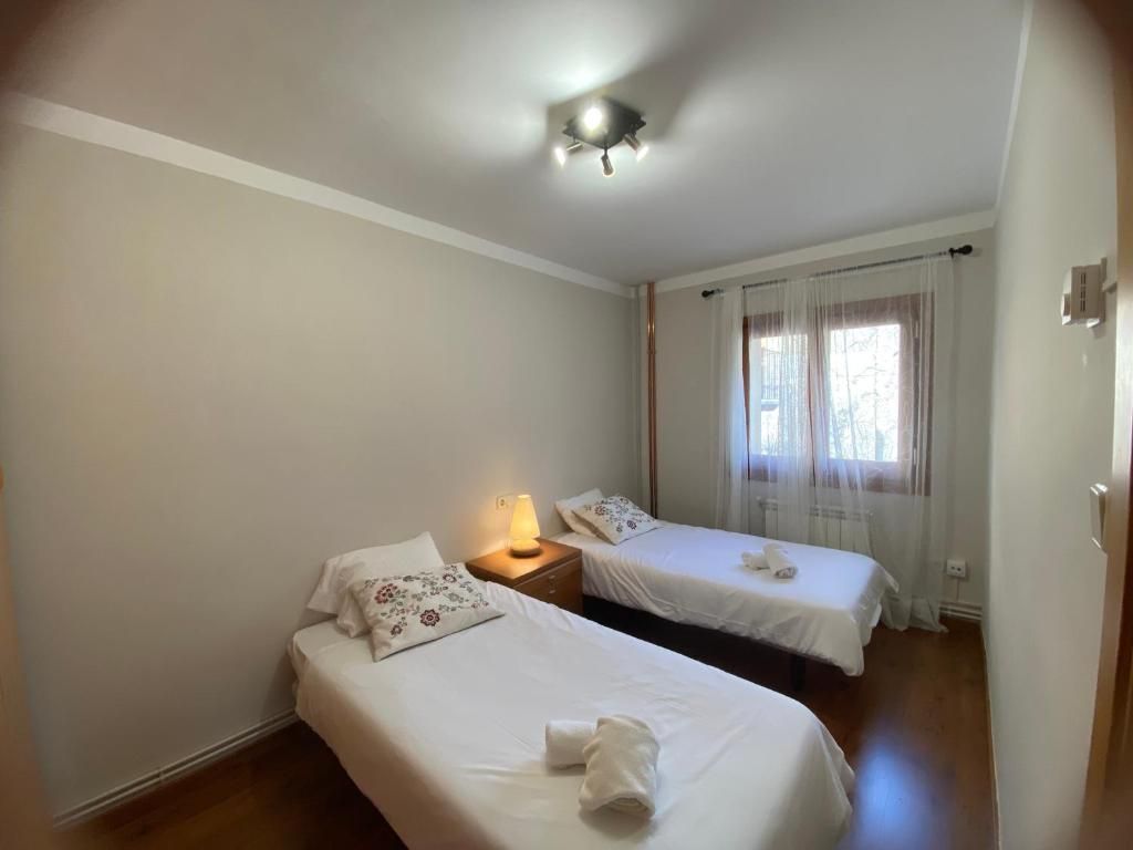 Dos llits individuals en una habitació amb finestra, tauleta de nit i llum de sostre. Roba de llit blanca.