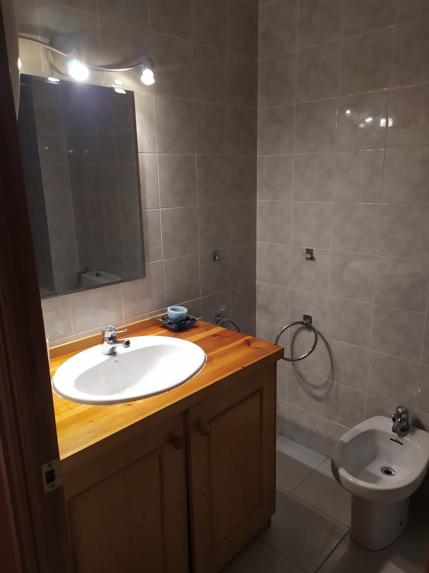 Bany amb tocador de fusta, pica blanca i bidet. Les parets estan enrajolades en un color neutre.