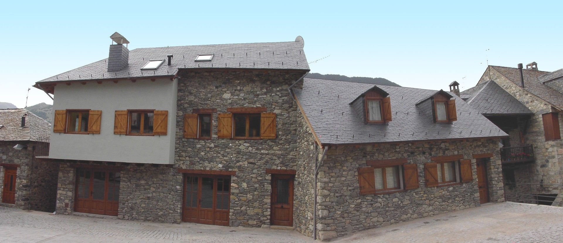 Casa de pedra amb persianes i porta de fusta, contra un turó i cel blau.