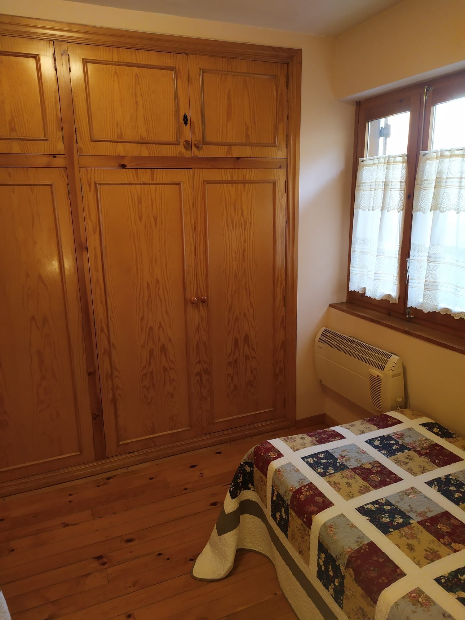 Dormitori amb armari de fusta, llit amb edredó, finestra petita i estufa de paret.