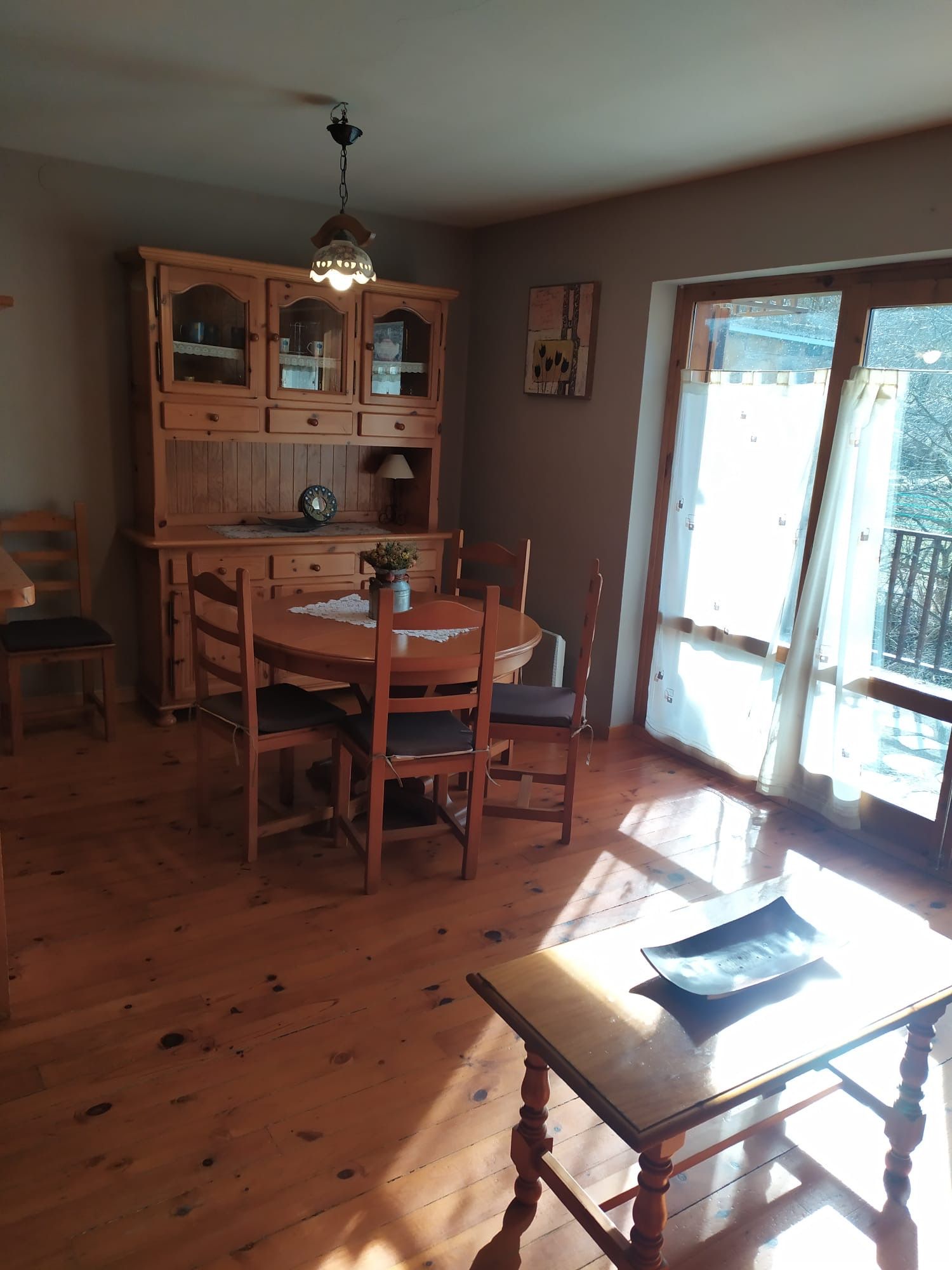 Menjador amb mobles de fusta: aparador, taula, cadires. La llum del sol entra a través d'una porta de vidre.