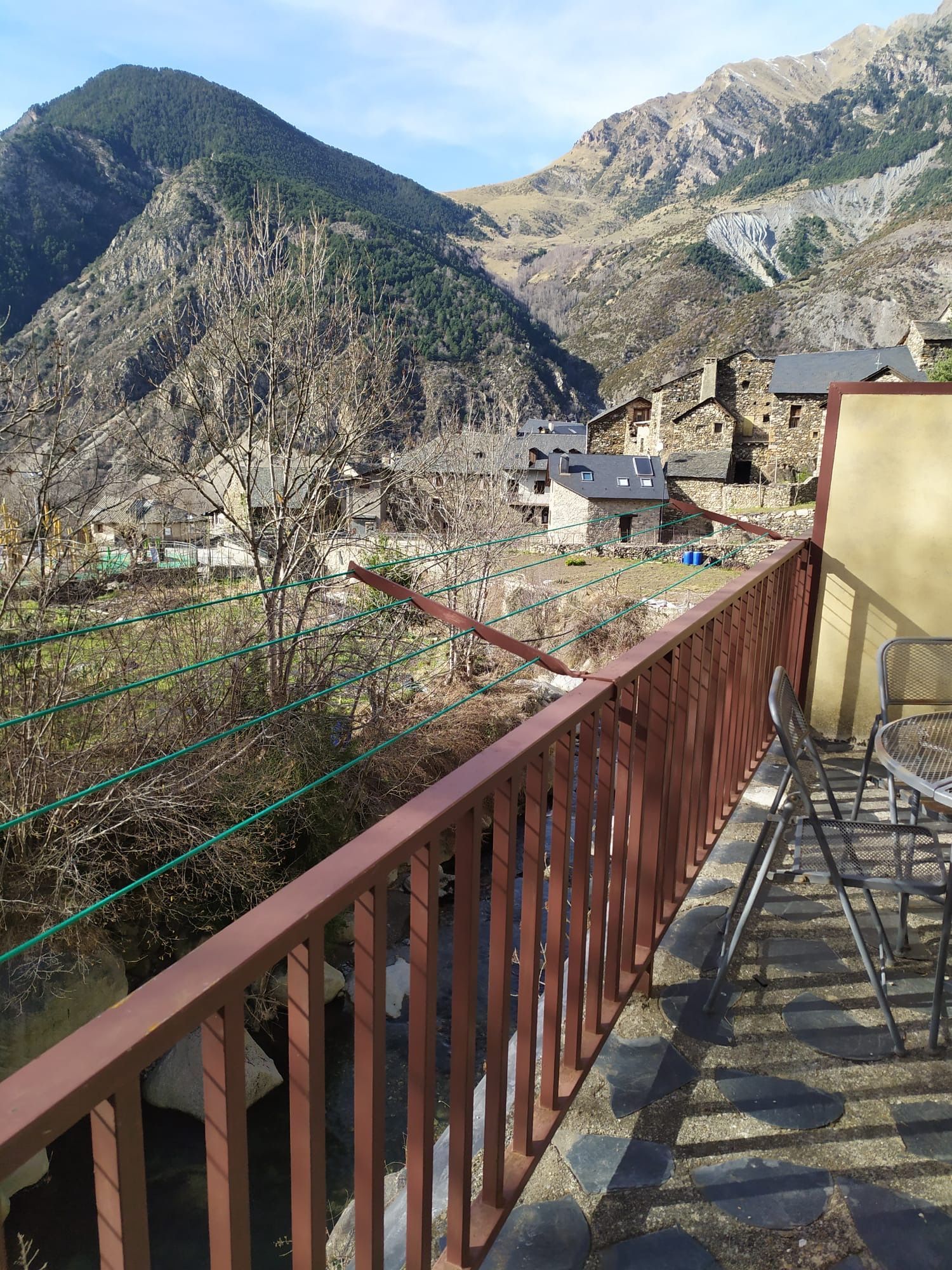 Vista des del balcó d'un poble enclavat en una vall amb muntanyes al fons. Barana i taula marrons en primer pla.