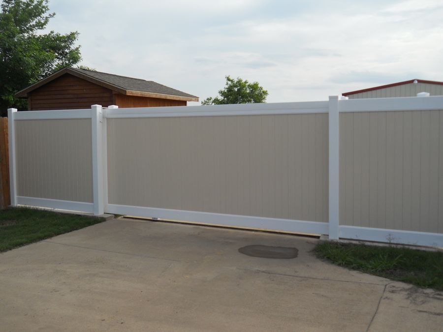 A white fence surrounds a driveway with a house in the background