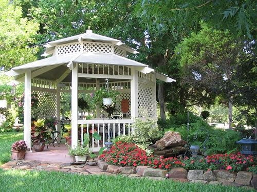 A white gazebo is surrounded by trees and flowers in a garden.