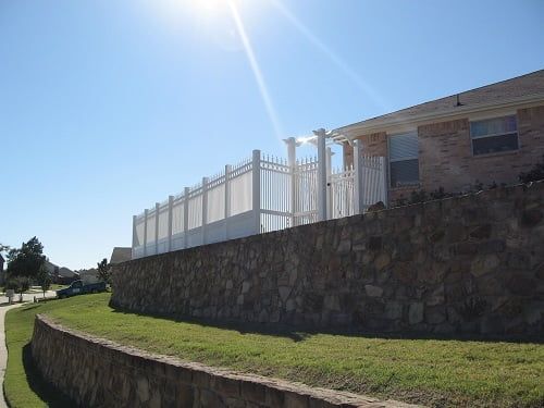A white fence is sitting on top of a stone wall next to a house.