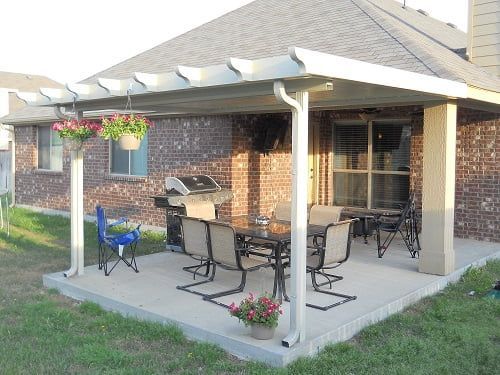 A patio with a table and chairs under a pergola in front of a brick house.