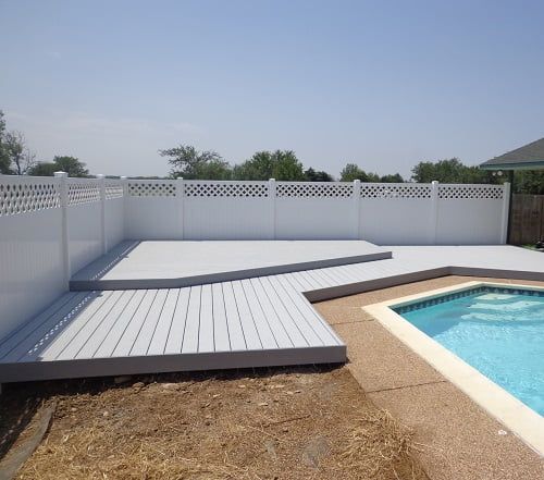 A white fence surrounds a swimming pool with a white deck in front of it.