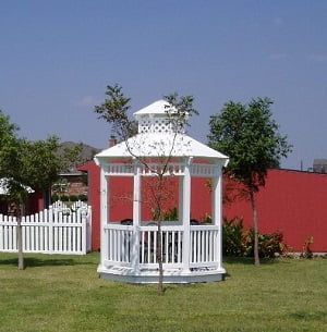 A white gazebo with a white picket fence in front of a red building
