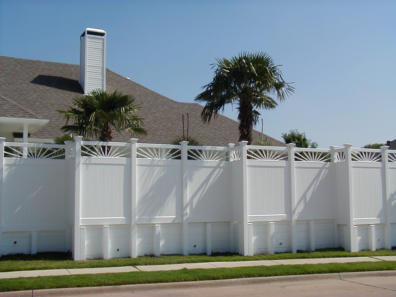 A white fence surrounds a house with palm trees in the background
