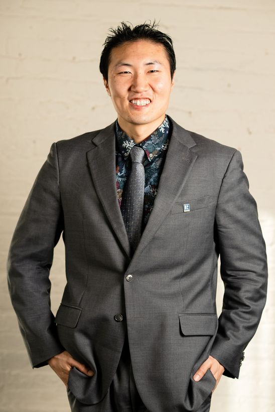 A happy man is standing at a gorgeous background with a gray suit on and a formal flower dress shirt.