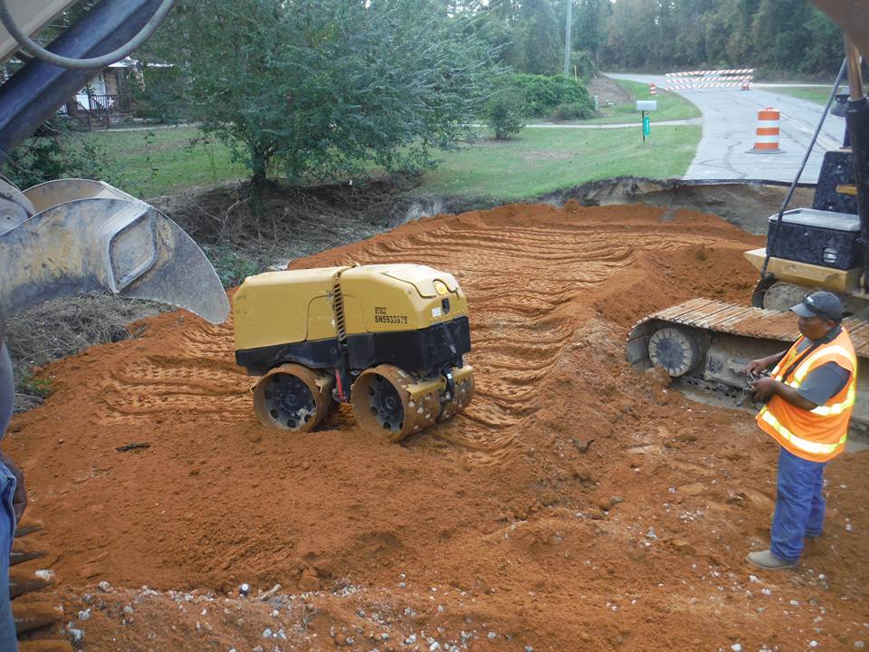 a man in an orange vest is standing in the dirt next to a bulldozer and a roller .