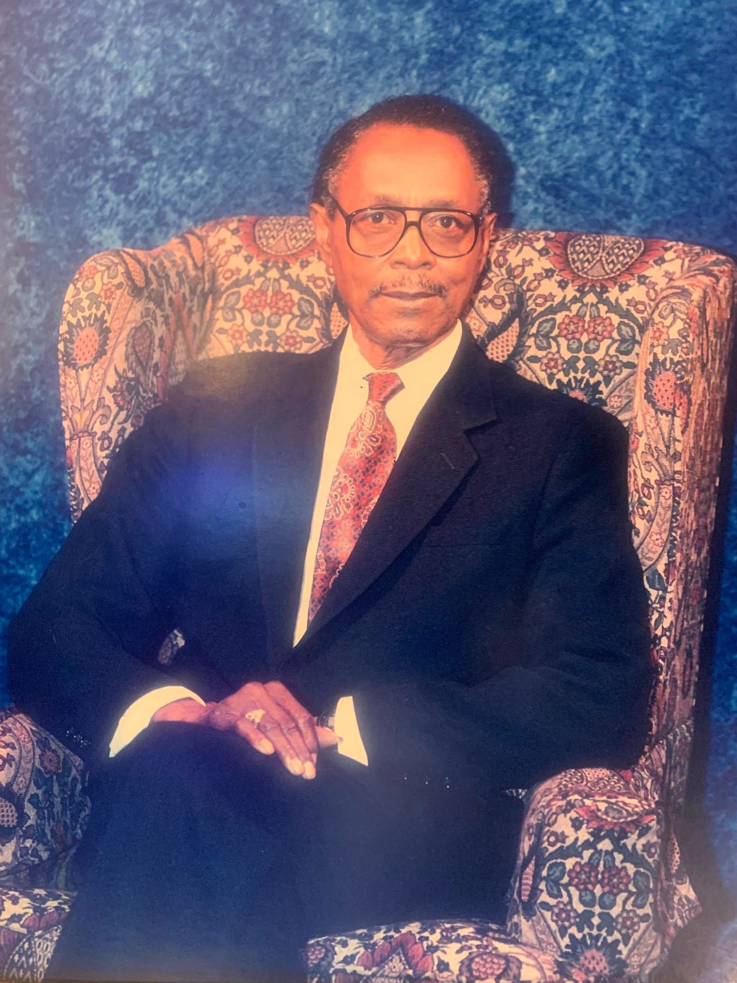 Man in suit seated in patterned armchair, blue backdrop.