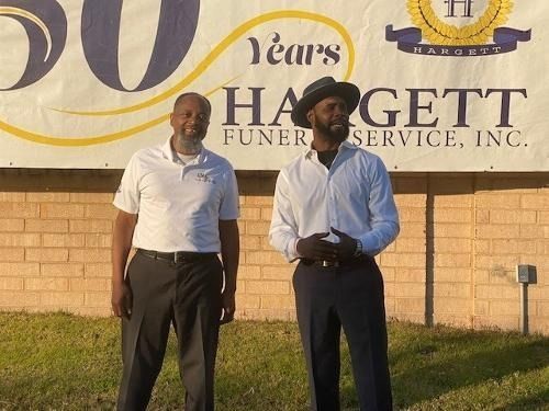 Two men stand in front of a banner for Hagett Funeral Service, Inc., celebrating 50 years.