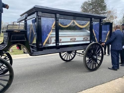 Black horse-drawn hearse with casket at a cemetery; men in suits stand nearby.