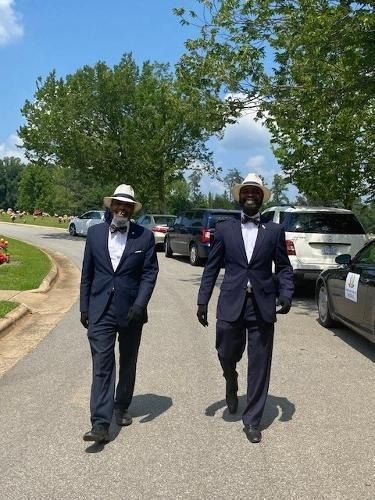 Two men in suits and hats walk on a road in a cemetery on a sunny day.