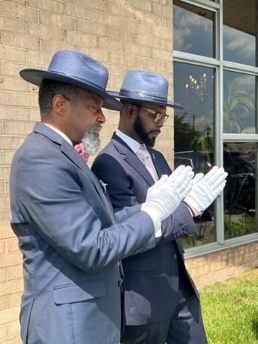 Two men in suits and hats with white gloves, praying outside a building.