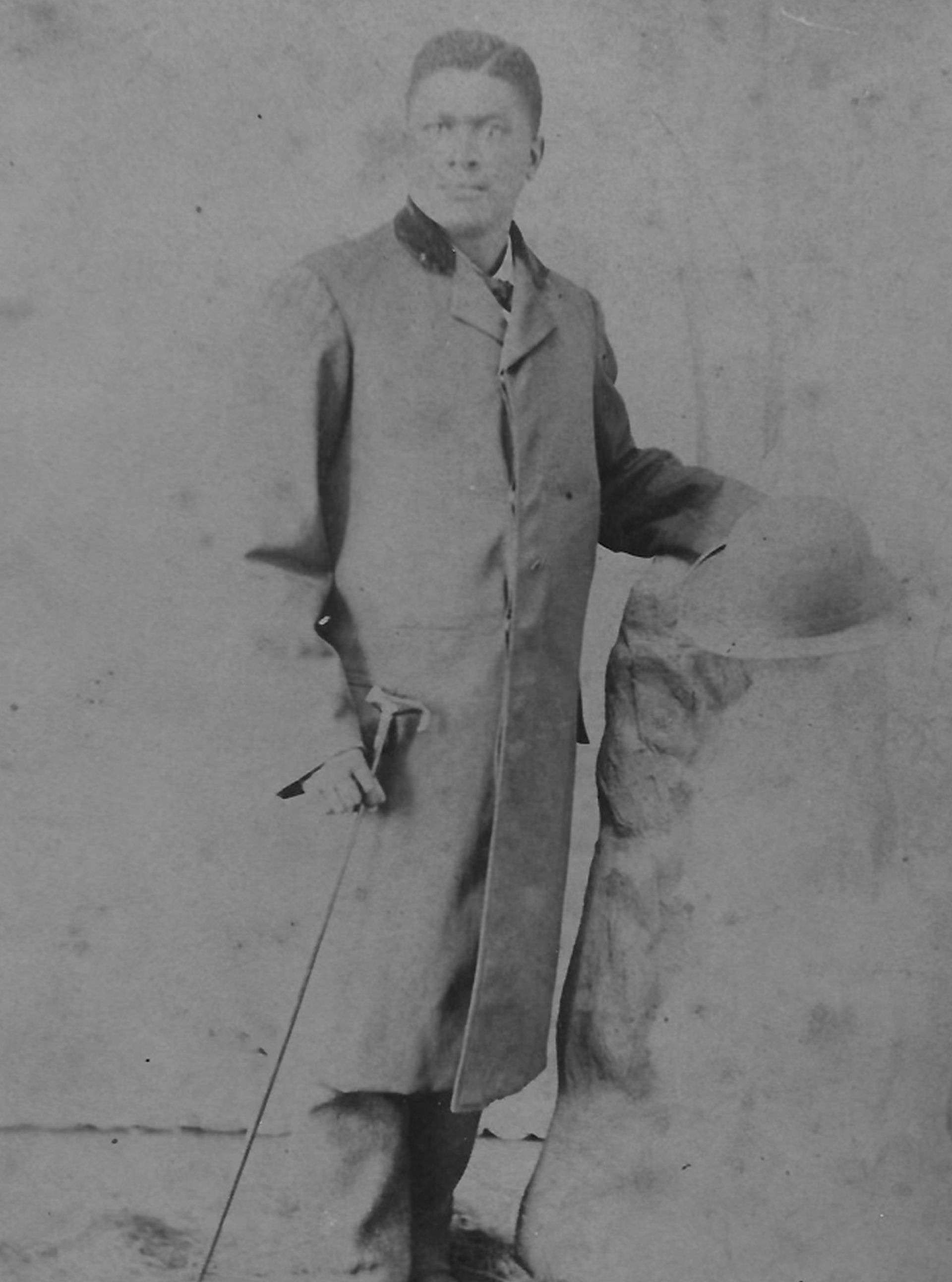 Man in long coat holds hat and cane, standing.