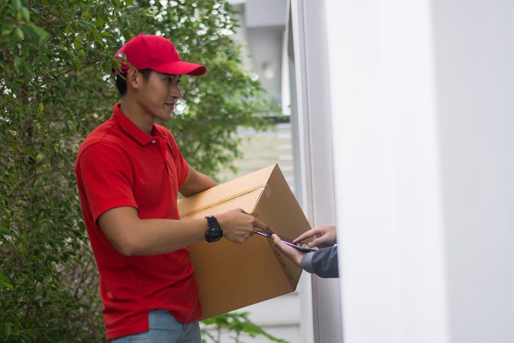 Un repartidor con camisa roja y gorra entrega una caja de cartón a una persona en una puerta.