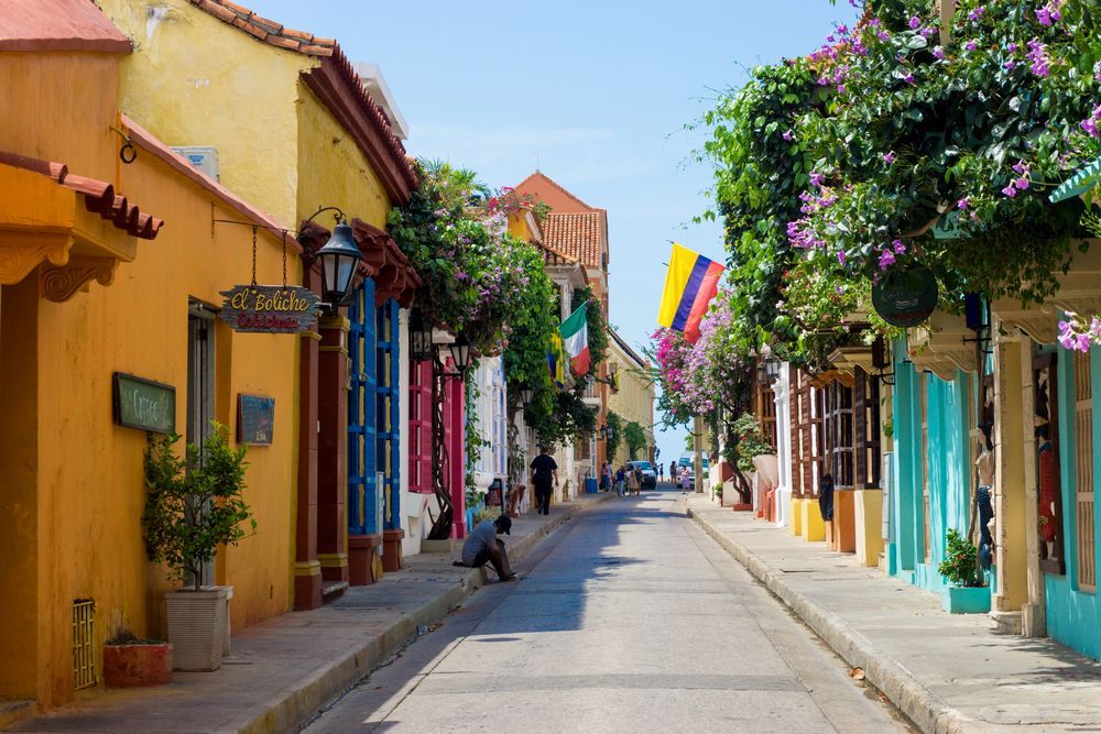 Calle colorida en Cartagena, Colombia, con edificios en amarillo, azul y rosa, bandera colombiana ondeando.