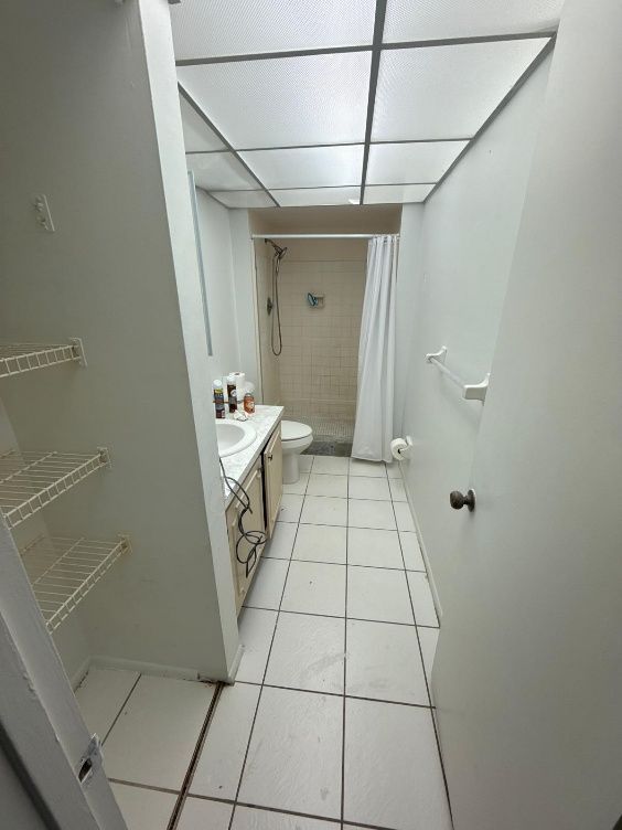 Narrow bathroom with white tile floor, toilet, vanity, and shower. Wire shelving to the left.