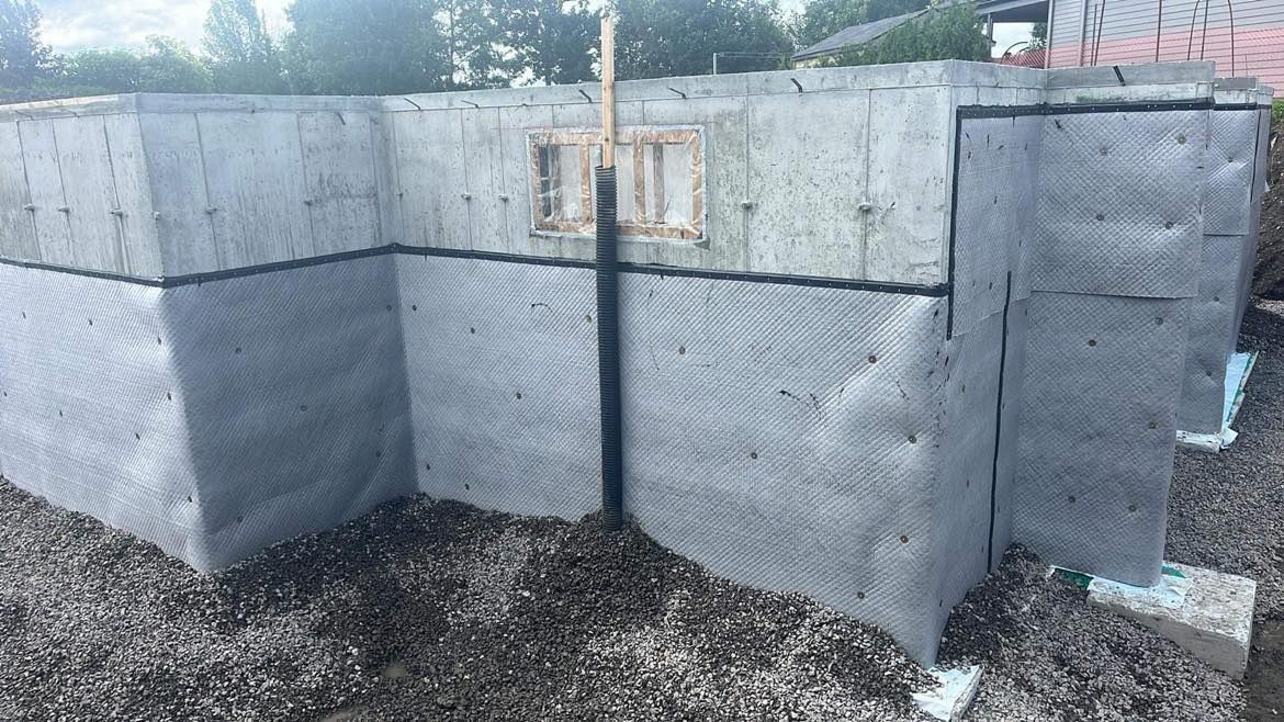 Fondations d'un bâtiment avec bâche de protection, une petite fenêtre et du gravier tout autour.