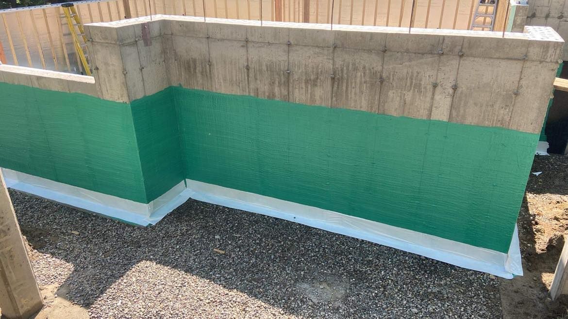 Mur de fondation en béton, membrane d'étanchéité verte, ruban blanc à la base, lit de gravier.