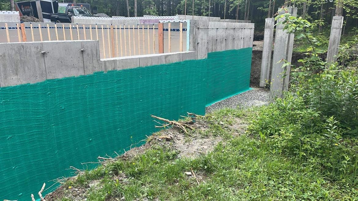 Fondations en béton avec membrane de drainage verte, base de gravier et ossature en bois.