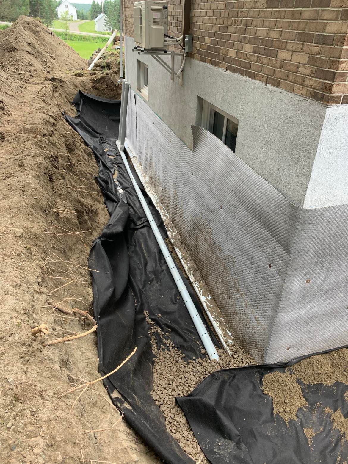 Tranchée creusée autour des fondations d'un bâtiment ; tuyau de drainage et gravier recouverts d'une bâche noire.