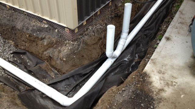 Tranchée avec tuyaux de drainage en PVC blanc, contre un bâtiment, sur une toile de paillage noire.
