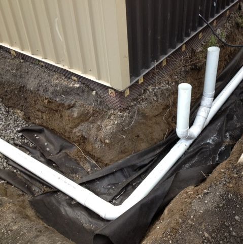 Des tuyaux en PVC blanc dans une tranchée à côté d'un bâtiment, laissant apparaître des tissus noirs et de la saleté.