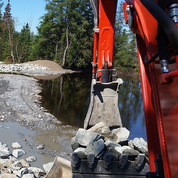 Godet orange d'une excavatrice ramassant des pierres d'une rive boueuse pour les déverser dans une étendue d'eau sombre ; forêt en arrière-plan.