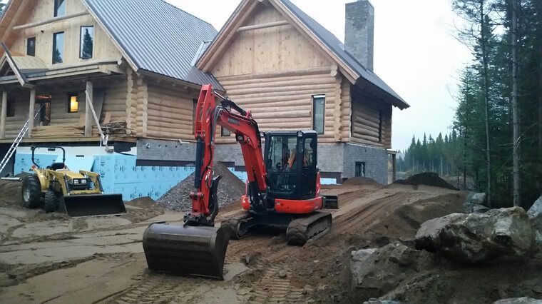 Une excavatrice et un tracteur travaillent sur un chantier de construction près d'une cabane en bois.