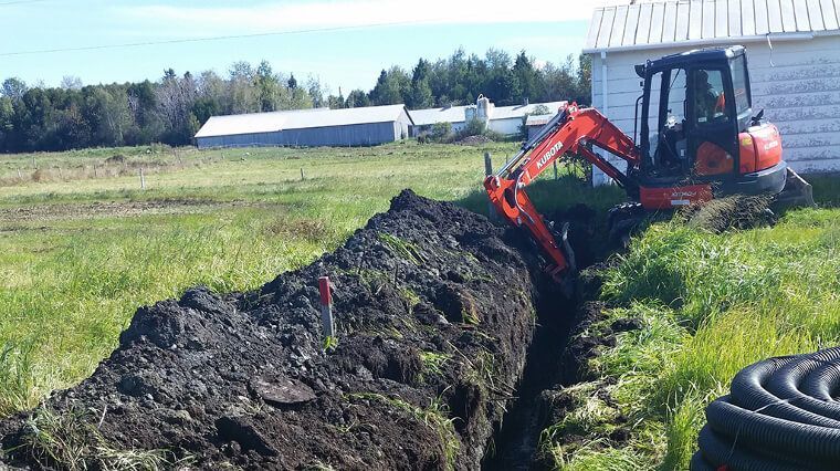 Une mini-pelle creuse une tranchée dans un champ herbeux à côté d'un bâtiment blanc.