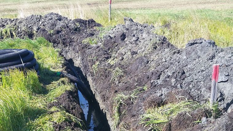 Tranchée creusée dans un champ, la terre s'y est accumulée sur les bords. Un tuyau de drainage noir se trouve à proximité.