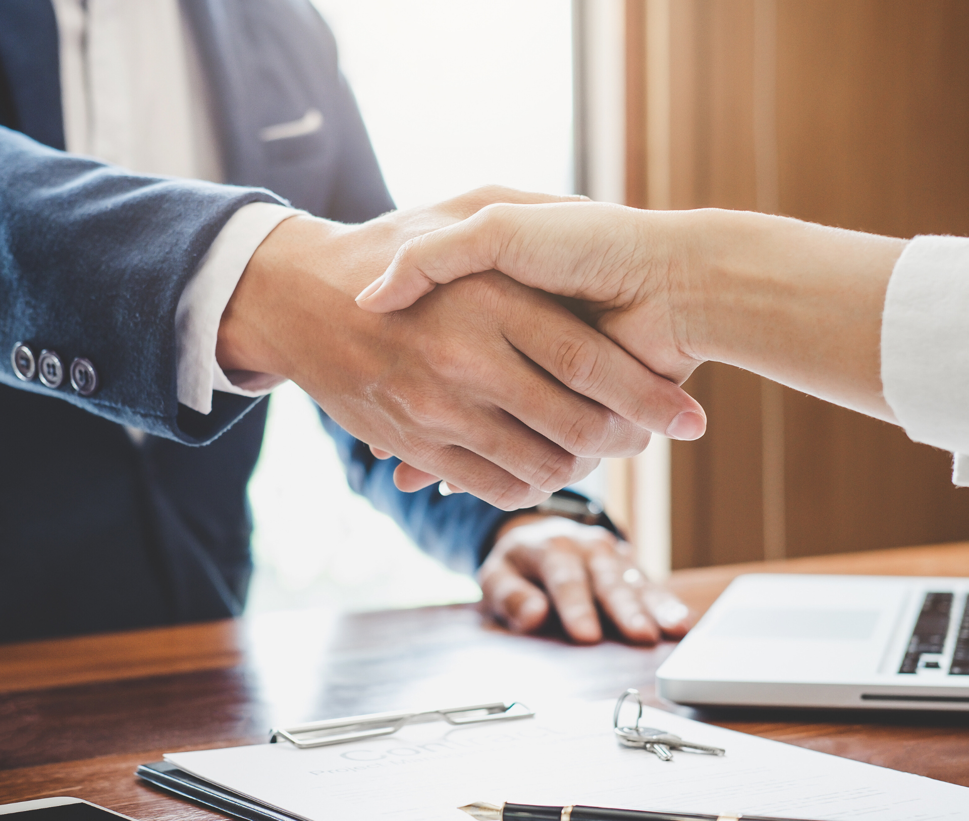 Two people shaking hands over a table with papers and a laptop.