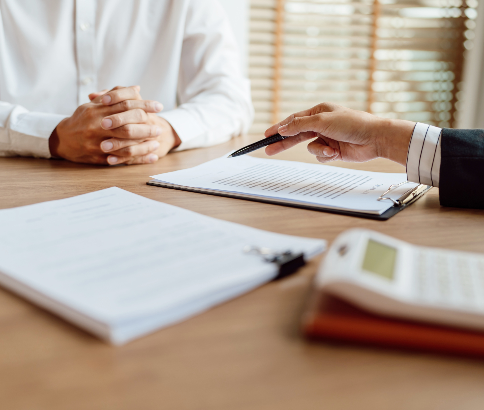 Person reviewing paperwork with another person seated across a desk. A hand points at the document.