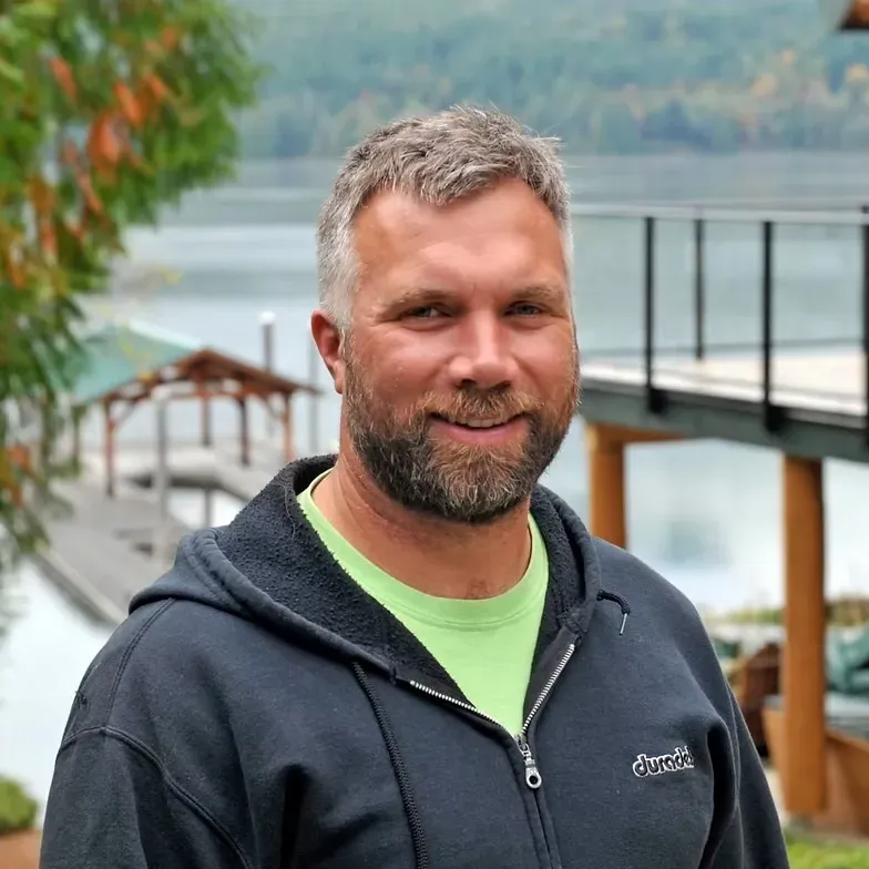 Man with graying hair and beard smiles, outdoors by a lake, wearing a hooded sweatshirt and light green shirt.