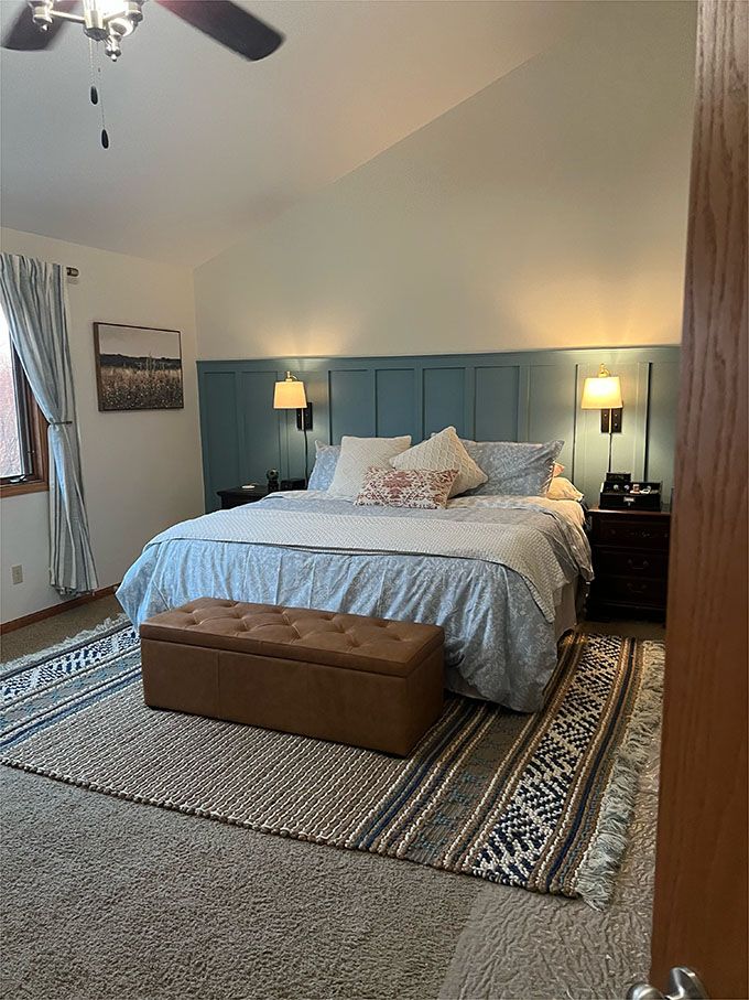 A bedroom with a king size bed , rug , ottoman and ceiling fan.