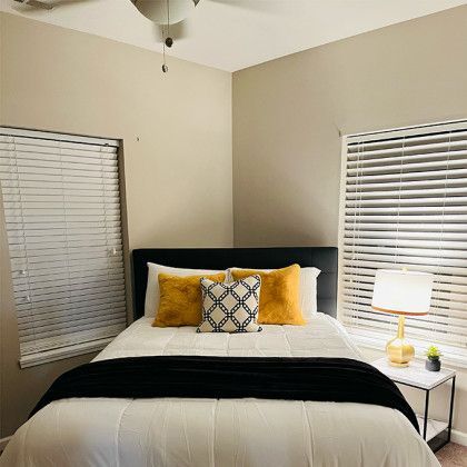 A bedroom with a bed , two windows , a lamp and blinds.